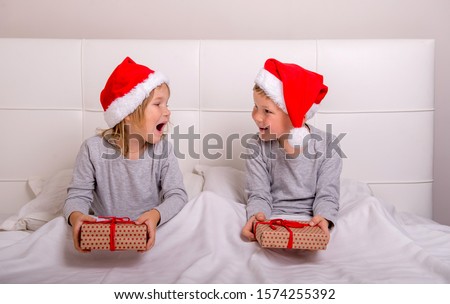 Funny couple boy and girl having fun in bedroom on Christmas morning. Kids in Santa Clause red hats and gifts in hands. Happy xmas and New Year. Children lying in bed and hiding under white blanket.
