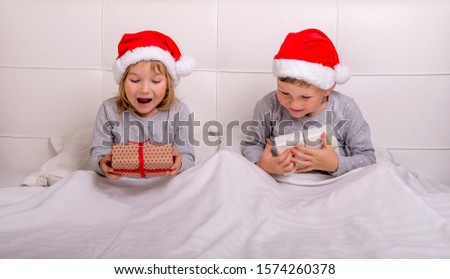 Funny couple boy and girl having fun in bedroom on Christmas morning. Kids in Santa Clause red hats and gifts in hands. Happy xmas and New Year. Children lying in bed and hiding under white blanket.