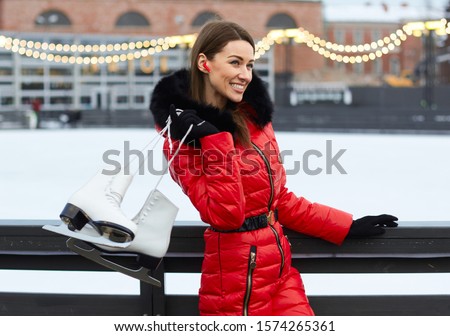 Young smile face brunette woman in red winter overall clothes stands at open air outdoor ice rink arena board fence with wireless earphones in ear and holds up by hand to fix hang skates on shoulder Royalty-Free Stock Photo #1574265361