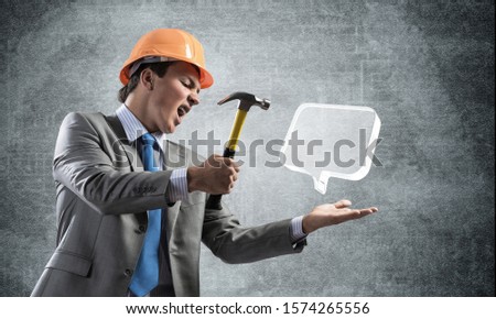Furious businessman going to crash glass speech bubble with hammer. Young handsome man in business suit and safety helmet standing on wall background. Business communication and online dialogue