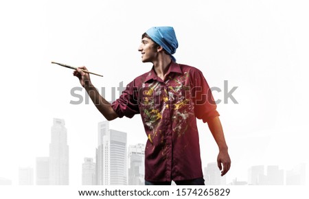 Young male artist holding paintbrush. Happy painter in dirty shirt and bandana standing on background modern cityscape. Creative hobby and artistic occupation concept with copy space