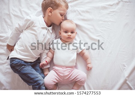 Kid boy 2-3 year old kissing baby girl 1 year old wearing casual clothes lying in bed on white linen in bed close up. Sister and brother. Childhood. 