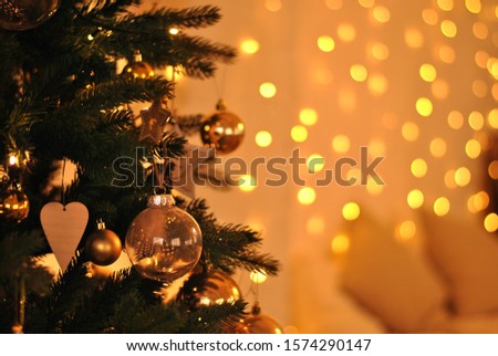 Very bright lights bokeh background lightened by golden yellow garland. Christmas fir tree decorated with balls and hears decorations on the front left side. Shiny beautiful new year holiday picture