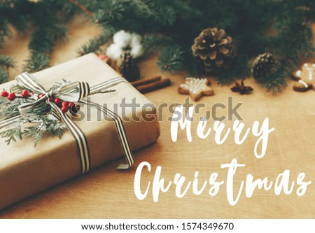 Merry Christmas text sign on stylish rustic christmas gift, pine branches, cones, gingerbread cookies, cinnamon, anise on rural wooden table, Season's greeting card