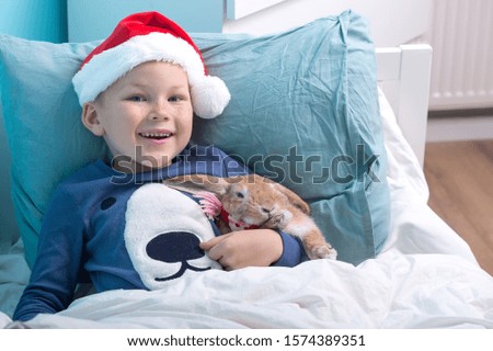 Funny child boy in Santa Clause hat and pajamas waking up with pet rabbit in Christmas morning. Happy child hugging his Christmas gift. Happy Xmas and New Year holiday! 