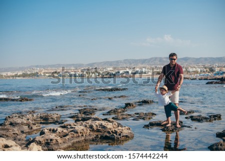 Father with a small son traveling near the sea. Dad and baby play on the stones on the beach against the background of the sea and the city on the horizon.