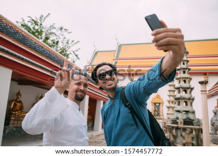Men in a Buddhist temple take selfies on the phone. Young guys in simple national t-shirts against the background of the sights of the palace make photos with a smartphone.