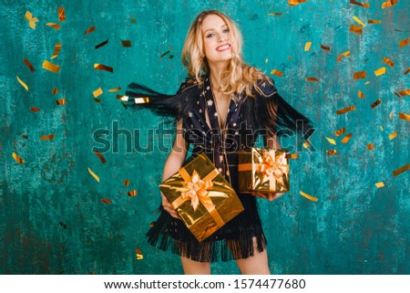 attractive happy smiling woman in stylish black dress with fringe dancing, celebrating christmas and new year with gifts, holding present box, golden confetti flying, party style, having fun