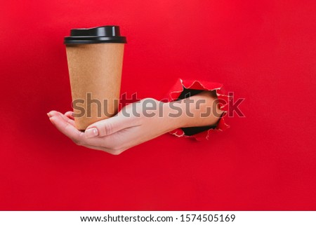 Female hand holding craft paper coffee cup through a hole in red background. Minimal mockup concept.