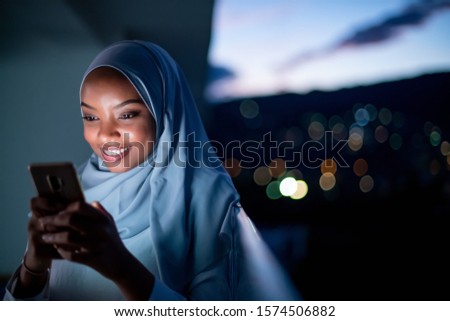 Young modern Muslim woman wearing scarf veil on urban city  street at night texting on smartphone with bokeh city light in background