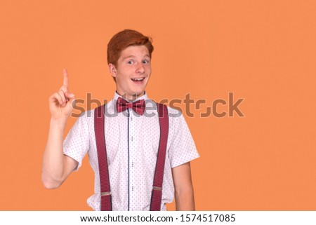 Half length  portrait of a   red-haired young smiling boy teenager wearing  white shirt with a red bow tie and suspenders  proving something by raising his index finger up against  orange background 