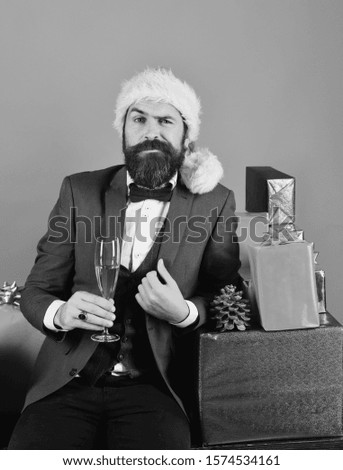Xmas corporate party concept. Man with beard holds champagne. Businessman with confident face near stack of boxes and glass of drink. Santa in retro suit by blue and red gifts on red background.