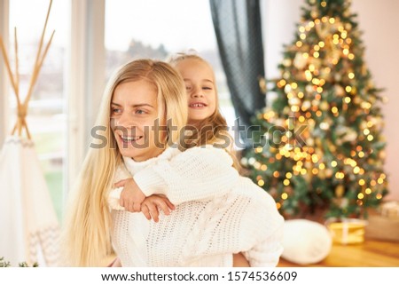 Indoor shot of happy young woman with long hair giving piggy back ride to her adorable little daughter, having fun, fooling around in living room with decorated shining New Year's tree in background
