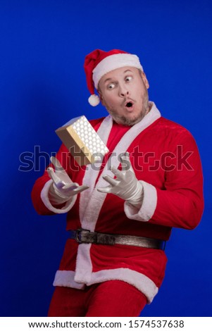 Emotional male actor in a costume of Santa Claus holds a box with a gift in his hands, juggles and poses on a blue background