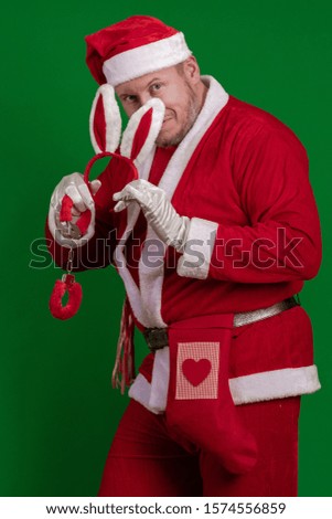 Santa Claus holds red fur handcuffs and a whip in his hands and poses on a green chrome background