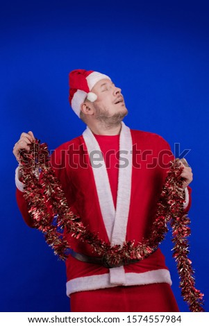 Male actor in a suit and hat of Santa Claus with a red tinsel garland dancing and posing on a blue background