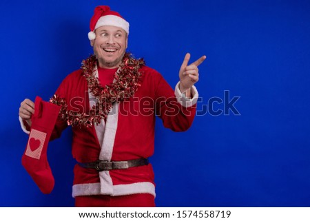 Emotional male actor in a costume of Santa Claus holds a Christmas sock in his hands with gifts and poses on a blue background