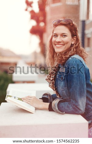 Young redhead Caucasian woman holding book outdoors at autumn, toothy smile, looking into camera