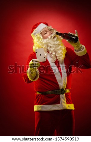 Male actor in a costume of Santa Claus holds playing cards and a bottle with alcohol in his hands, drinks and poses on a dark red background