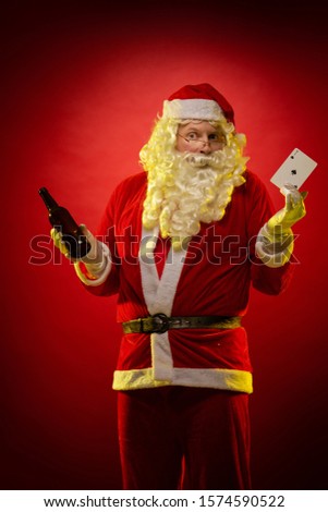 Male actor in a costume of Santa Claus holds playing cards and a bottle with alcohol in his hands, drinks and poses on a dark red background