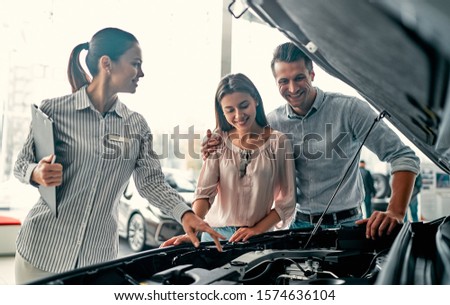 Buying their first car together. Young car saleswoman standing at the dealership telling about the features of the car under the hood to the customers.