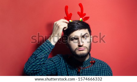 Portrait of amused young man, wearing green shirt, deer horns and Christmas jewelry, isolated on red background.