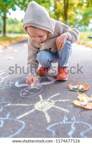 Little boy in autumn on street, draws on pavement, multicolored crayons. Warm clothes on a cold day, beige cardigan with a hood. Weekend breaks, creativity and drawings