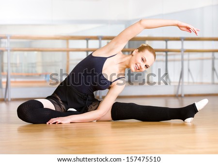 Bending ballet dancer stretches herself on the wooden floor in the classroom