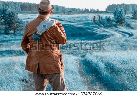 a man with a brown suit and standing back on shoulder gas mask in the background of blue grass. the grass looks a funny colour postapokalipsis