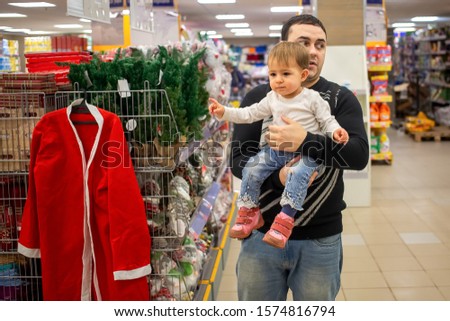 A young Caucasian man holds a small cute baby in his arms in a store near a bookcase. baby points with finger at Christmas Santa costume and New Year decorations. close-up, soft focus, blur background