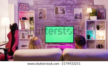 Young couple enjoying playing online games together sitting on sofa. Tv with green screen.