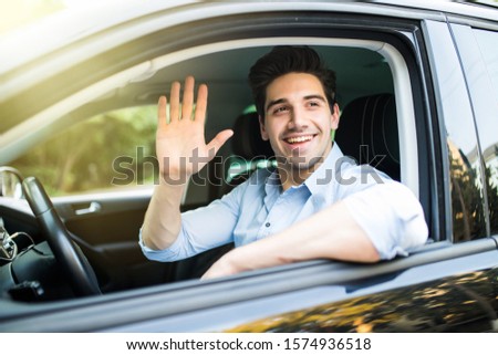 Portrait of young attractive handsome brunette man driving car and greeting somebody with hand. Royalty-Free Stock Photo #1574936518