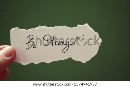 Hand holding piece of paper note with text "Be Strong" closeup blur green background