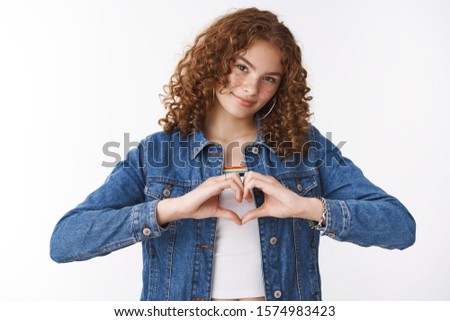 Friendship, relationship lifestyle concept. Charming friendly european redhead curly-haired girl show love sign make heart near chest tilting head smiling expressing sympathy, white background
