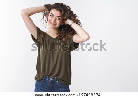 Joyful young carefree armenian girl playing curly hair comb, hold hands behind smiling joyfully making hairstyle near mirror, standing casually white background wearing olive t-shirt jeans