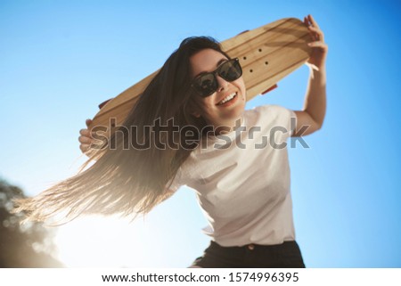 Low-angle shot cheerful happy young brunette woman enjoy riding skateboard, skater girl spend weekends skate park, hold wooden penny board behind head, laughing look down at camera