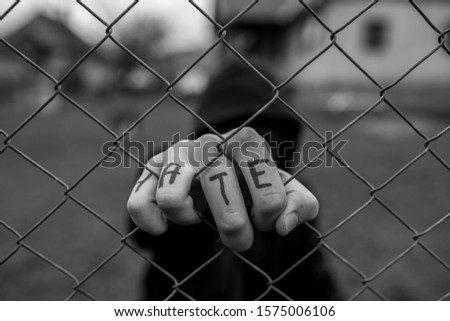 Aggressive teenage boy holding the wired fence at the correctional institute, the word hate is written on hes hand, conceptual image of juvenile delinquency in black and white. Royalty-Free Stock Photo #1575006106