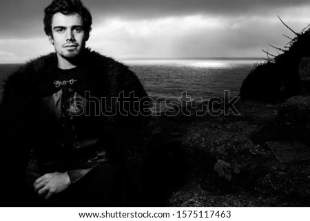 Portrait of handsome knight with goatee and fur coat looking at camera with ocean and spot in background
