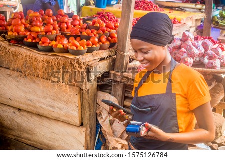 young african woman selling in a local african market holding a mobile point of sale system and using her mobile phone Royalty-Free Stock Photo #1575126784