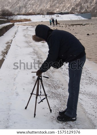 Photographer with tripod is taking picture of people and nature on the riverbank in winter.