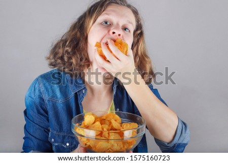 Portrait of a cute young girl with a glass bowl full of chips puts a lot of chips in her mouth.