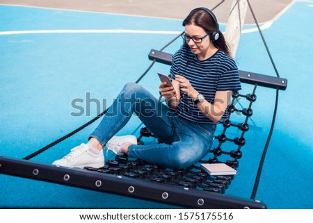 The girl, a brunette, pretty, with glasses, sits in a hammock, sways, on the playground with a bright blue coating, listening to music, dancing, relaxing, singing, trying to catch a wifi. Outdoor.
