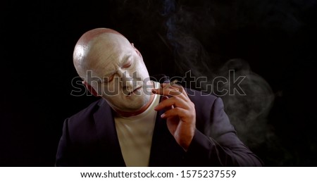 adult bald man with white greasepaint on his face is smoking cigarette in black background