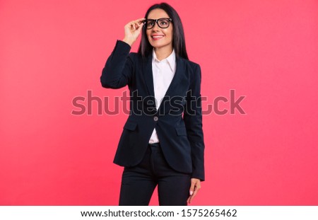 Smart and successful. Cute young business woman in smart outfit is posing on the pink background, holding her glasses with the fingers of her right hand, looking upwards and smiling.