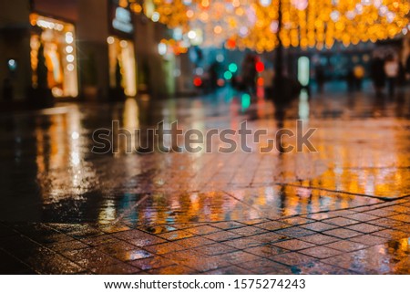 Rainy night in a big city, reflections of lights on the wet road surface. The view from the street level. Pedestrian feet and abstract background.