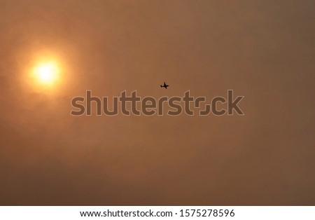 A firefighting aircraft flies high in the sky, as the sun glows through thick smoke; during widespread bushfires - and a state of emergency - across eastern Australia, in 2019.                      