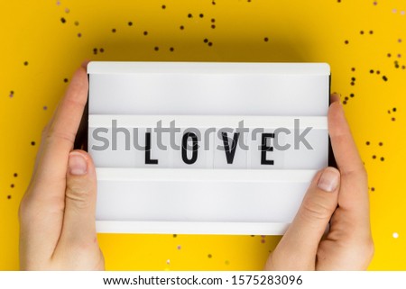 Love text on white lightbox. Yellow background with multicolored confetti. Flat lay style. Woman hand holding board