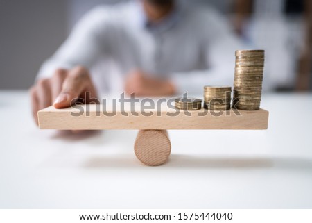 Close-up Of A Businessman Balancing The Coin Stack On Wooden Seesaw Over Desk Royalty-Free Stock Photo #1575444040