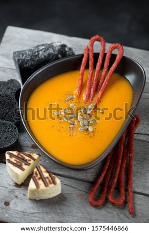 Bowl of pumpkin cream-soup with smoked meat, black baguette and grilled cheese, vertical shot on a grey wooden table