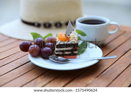 Blurred food background. On a wooden table is a white plate with a piece of chocolate cake, a spoon, a cup of coffee and a bunch of grapes. In the close-up view is a sun hat. Close-up, side view